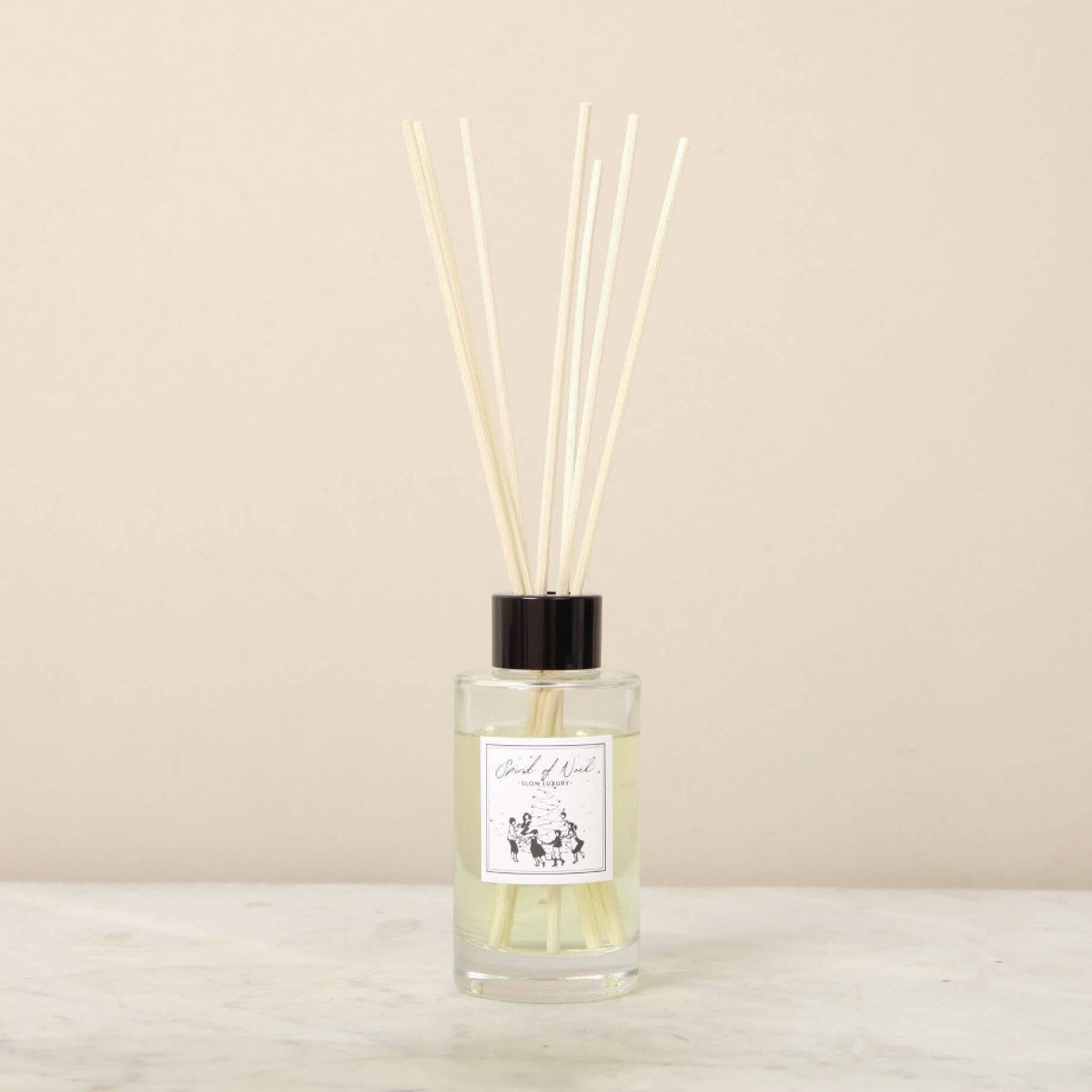 The Norfolk Natural Living Spirit Of Noel Reed Diffuser Set features a clear glass bottle with pale essential oils, a black cap, long thin reeds, and a white label with black text, displayed on a light surface against a beige background.