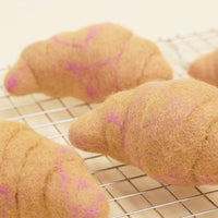 The MODERNBEAST WOOL CROISSANT Wool Dog Toy features three plush, wool-felt croissants in light brown and pinkish tones, displayed on a metal cooling rack against a pale background.
