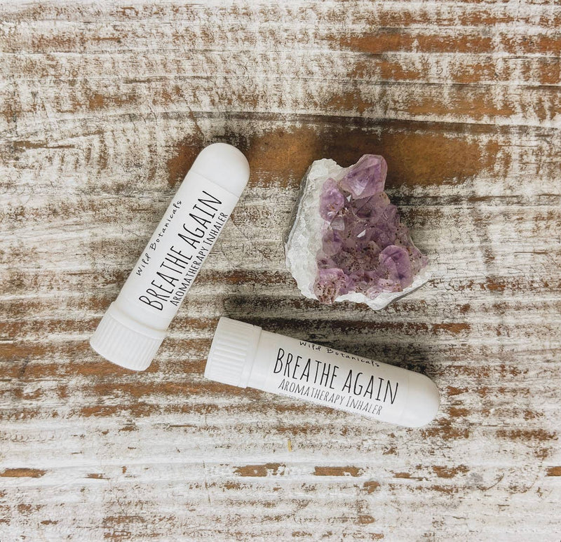 Two white Wild Botanicals AROMATHERAPY INHALERS lie on a rustic wooden surface alongside a small cluster of amethyst crystals. The essential oil-diffusing inhalers are positioned parallel to each other, with one slightly angled, promising to clear sinuses and rejuvenate your senses.