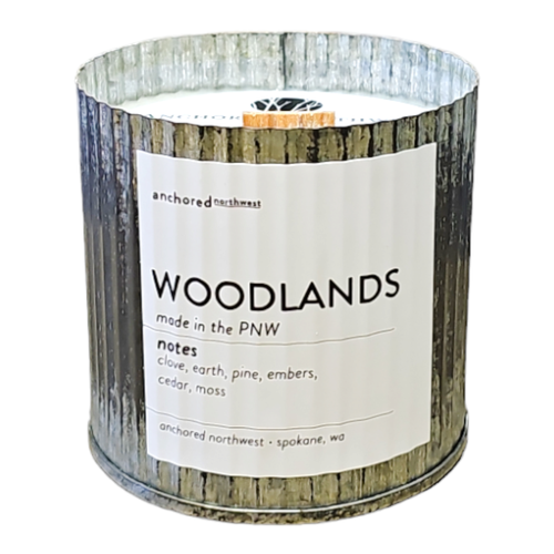 A rustic vintage candle made by Anchored Northwest, labelled "Woodlands," features a corrugated metal exterior. This soy wax candle exudes notes of clove, earth, pine, embers, cedar, and moss, embodying authentic charm from Spokane, WA.