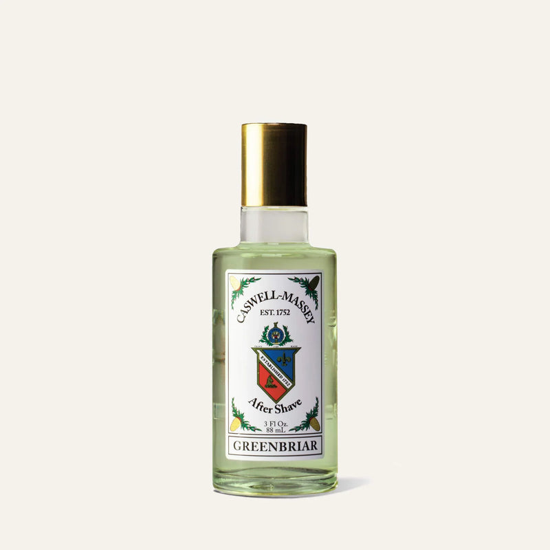 A clear glass bottle of Caswell-Massey Greenbriar Aftershave, a barbershop-inspired tonic with a gold cap and crest-decorated label featuring green accents and text detailing this essential Caswell Massey post-shave ritual.