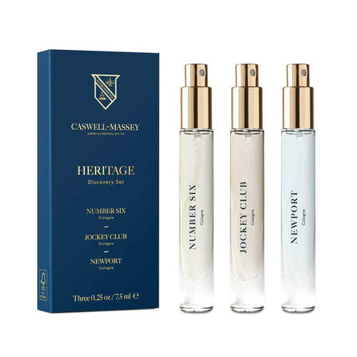 Three glass spray bottles, labeled "Number Six," "Jockey Club," and "Newport," are positioned beside a blue box with gold accents, titled "Caswell - Massey Heritage Cologne Discovery Set." This elegant set by Caswell Massey captures the essence of American history with its evocative fragrances and a logo featuring a shield design.