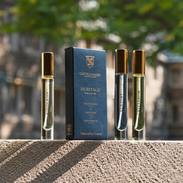 Three glass perfume vials with gold caps stand beside a Caswell-Massey Heritage Cologne Discovery Set box on a stone ledge, with blurred trees and buildings in the background—a classic gift for lovers of distinguished men's fragrances.