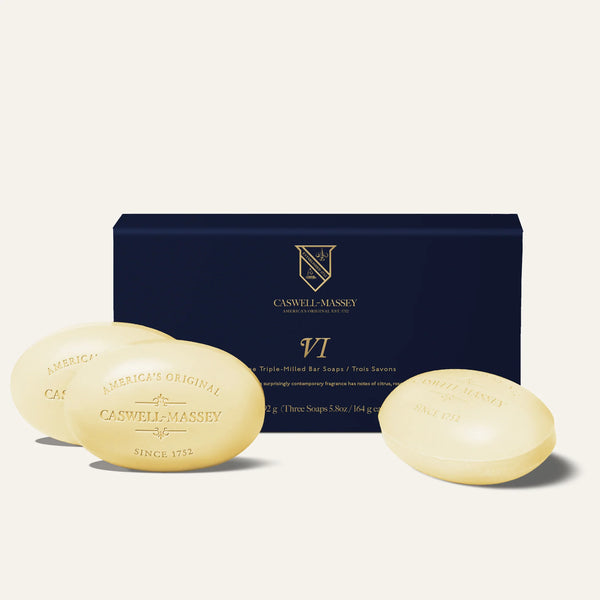 Three oval Caswell-Massey Number Six Bar Soaps, each triple-milled and embossed, are shown before a dark blue box labeled “VI” with gold branding. Both the box and soaps display gold details against an off-white background.