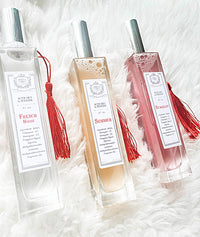 Three rectangular Z&Co. Farmhouse Collection Body Mist Perfume bottles with red tassels lie on a fluffy white surface, each providing an elegant, long-lasting scent and moisture for skin and hair. Labels read "French House," "Summer," and "Stardust.