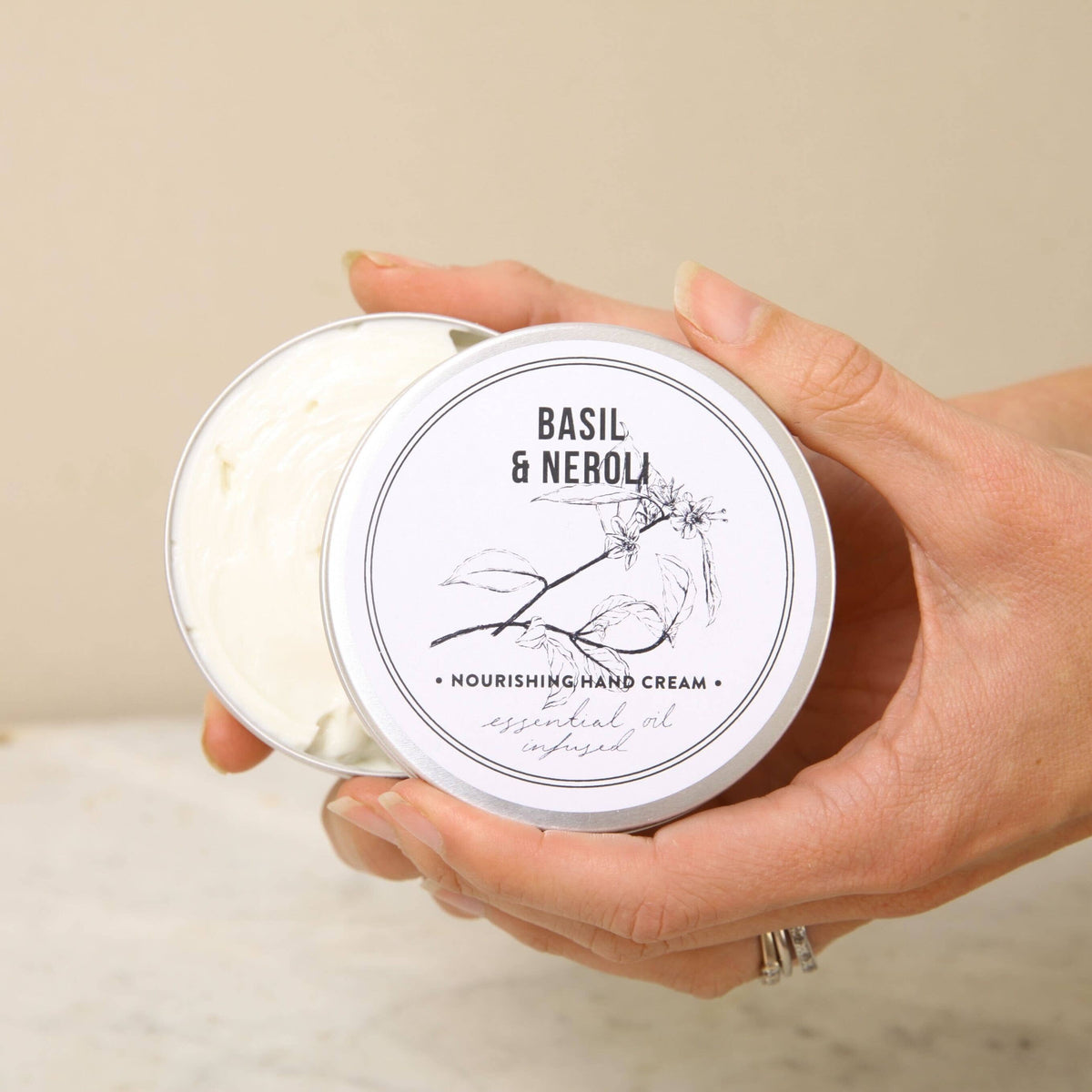 A person holds an open tin of Norfolk Natural Living Basil & Neroli Gardener's Hand Cream (100ml), revealing its creamy texture. The lid shows a botanical design and minimalist black branding.
