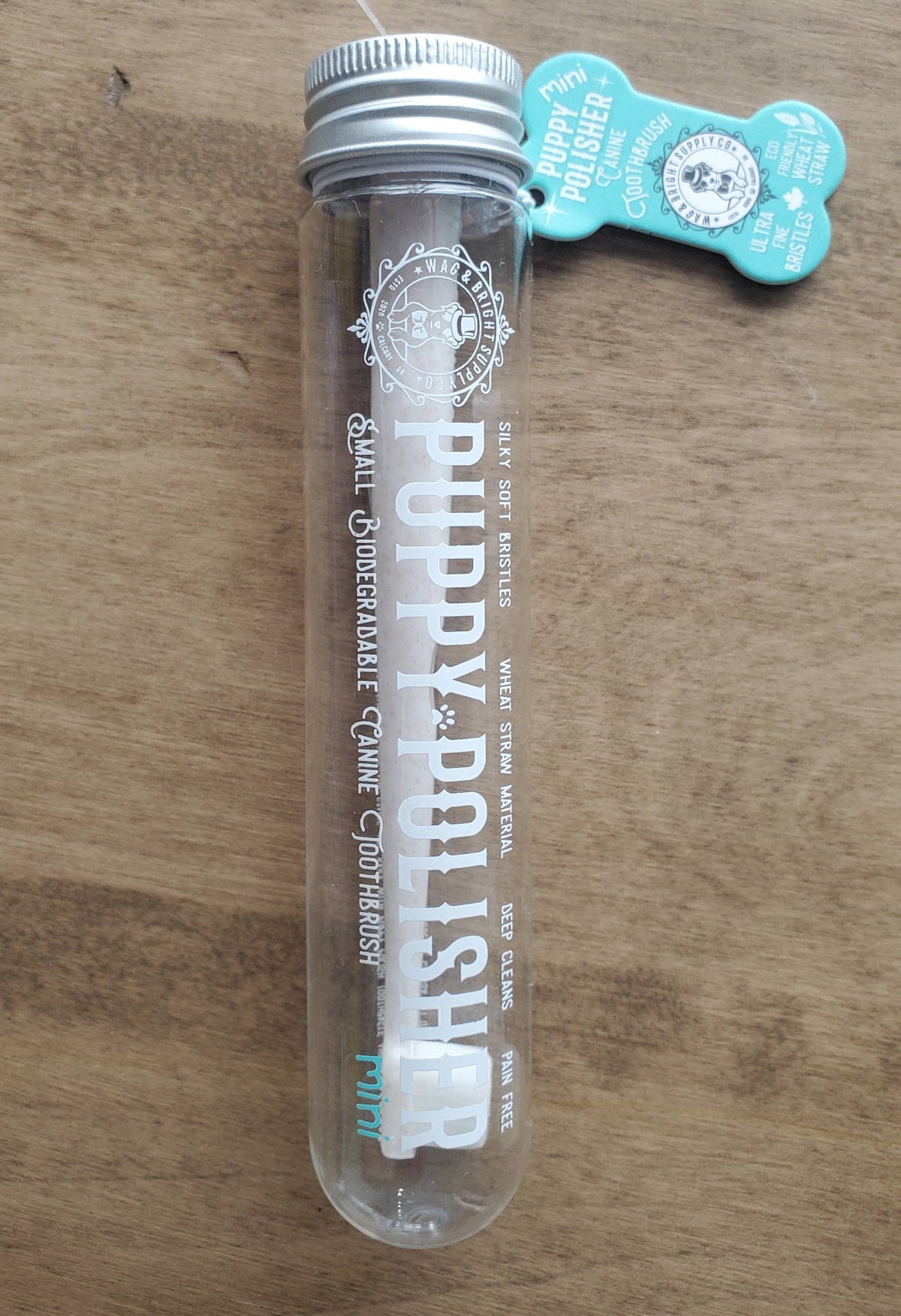 A clear tube labeled "Wag & Bright Supply Co. - Puppy Polisher Mini Eco Toothbrush (Small)" sits on a wooden surface. Made from sustainable bioplastic, it features a mint flavor seal and natural ingredients. The metallic cap secures the tube, while two tags hang near the top, ensuring it's an eco-friendly choice for your pup.