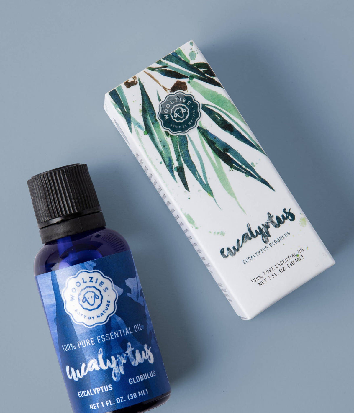 A blue bottle of Woolzies Eucalyptus Essential Oil sits next to its white, green-leaf adorned box on a light blue background; this 100% pure oil by Woolzies is known for supporting respiratory health.