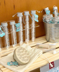 A display of pet grooming products features tubes of "Pup Polish" in test tube-like containers, a rubber grooming tool, and a small bucket filled with Wag & Bright Supply Co.'s Puppy Polisher Eco Toothbrush in medium size, crafted from wheat straw bioplastic—all set against a wooden background.