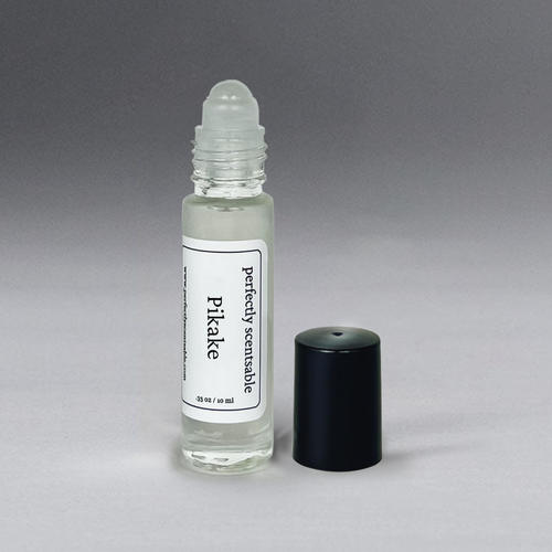 A small, transparent glass bottle with a rollerball applicator and a black screw cap, labeled "Perfectly Scentsable Pikake Perfume Oil - Rollerball." The bottle contains a clear liquid infused with a jasmine blend, and the cap is placed beside the bottle on a gradient gray background.