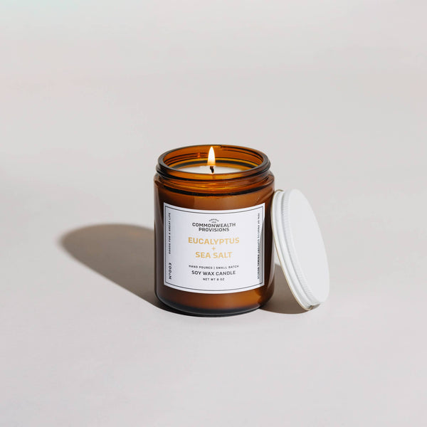 A lit Commonwealth Provisions Eucalyptus + Sea Salt Soy Candle in a brown glass jar with a white lid beside it, set on a neutral background—crafted with phthalate-free fragrance oils.