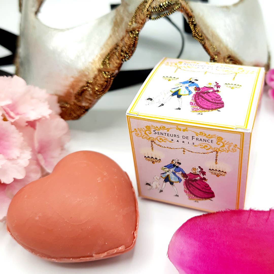 The Senteurs De France Red Fruit Heart Soap – Royal Masquerade Ball sits beside a decorative box with a dancing couple, pink flowers, and a gold-trimmed mask, capturing the elegance and romance of Versailles and French heritage.