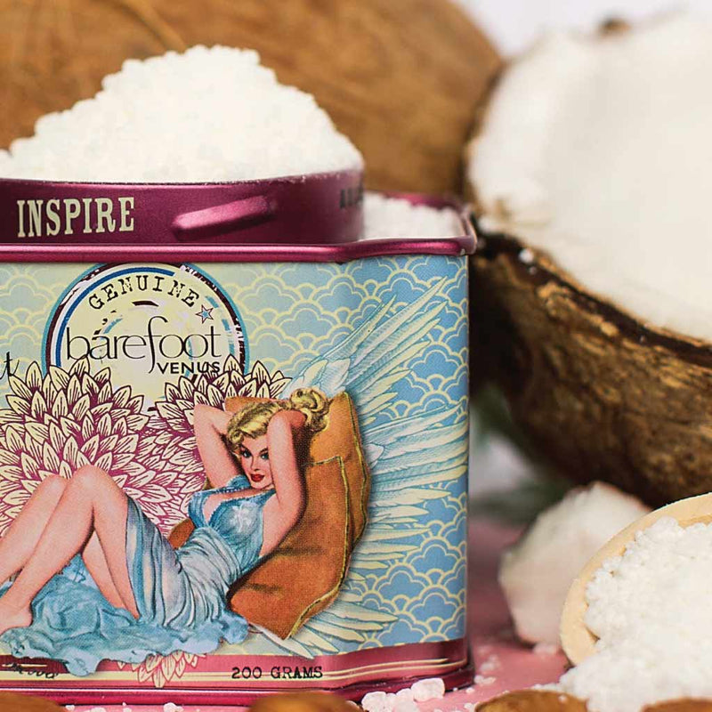 A vintage-style tin labeled "Barefoot Venus," featuring an illustration of a lounging woman with angel wings, rests surrounded by coarse white bath salts. The tin bears the word “INSPIRE” on its lid and a halved coconut is visible in the background. Enjoy your Barefoot Venus Coconut Kiss Bath Soak made with plant-based ingredients.