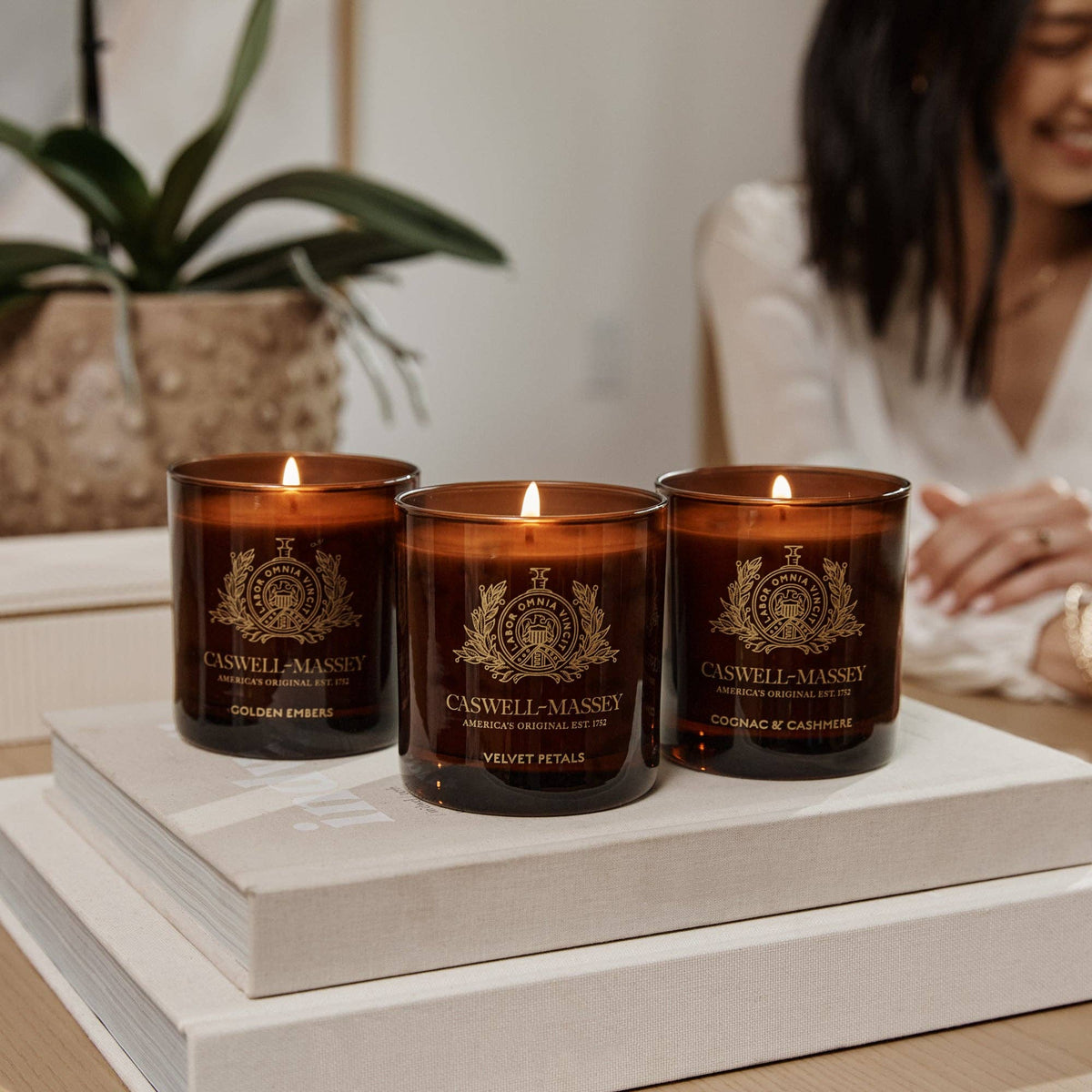 The Caswell-Massey Candle in Golden Embers burns atop stacked books, its meditative scent filling the room as a blurred figure sits nearby, close to a potted plant.