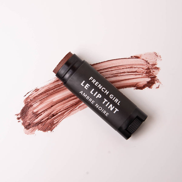 A tube of FRENCH GIRL Tinted Lip Balm in Ambre Noire, a nourishing tinted balm, sits on a white surface with a smear of its rich brownish-red shade behind it.