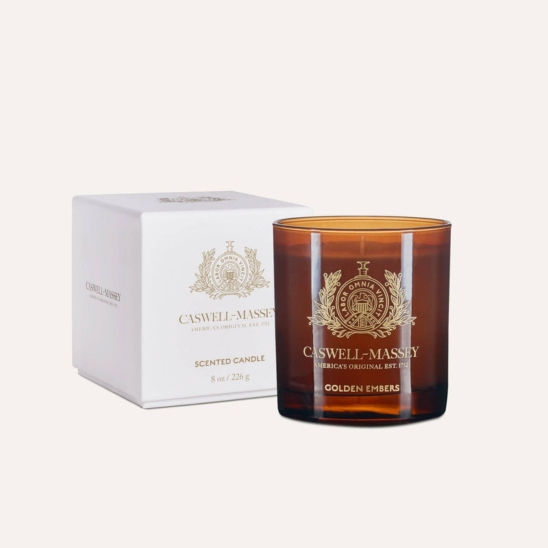 The Caswell-Massey Candle - Golden Embers, an 8 oz (226 g) hand-poured soy-blend candle in amber glass with a meditative scent, sits beside its white and gold box adorned with ornate crests and Caswell-Massey branding.