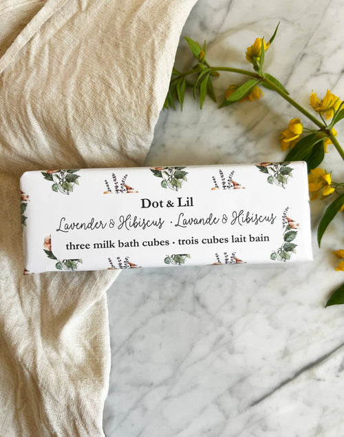A Dot & Lil Lavender & Hibiscus Milk Bath Cubes Trio Gift Set sits on marble, accented with a beige cloth and yellow flowers. The box, adorned with floral illustrations, displays both English and French text.