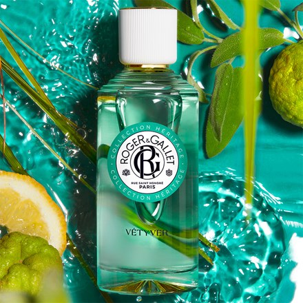 A bottle of Roger & Gallet Vetyver Wellbeing Fragrant Water - 3.3 oz, surrounded by water splashes, citrus slices, and green leaves on a vibrant turquoise background.