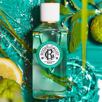 A bottle of Roger & Gallet Vetyver Wellbeing Fragrant Water - 3.3 oz, surrounded by water splashes, citrus slices, and green leaves on a vibrant turquoise background.