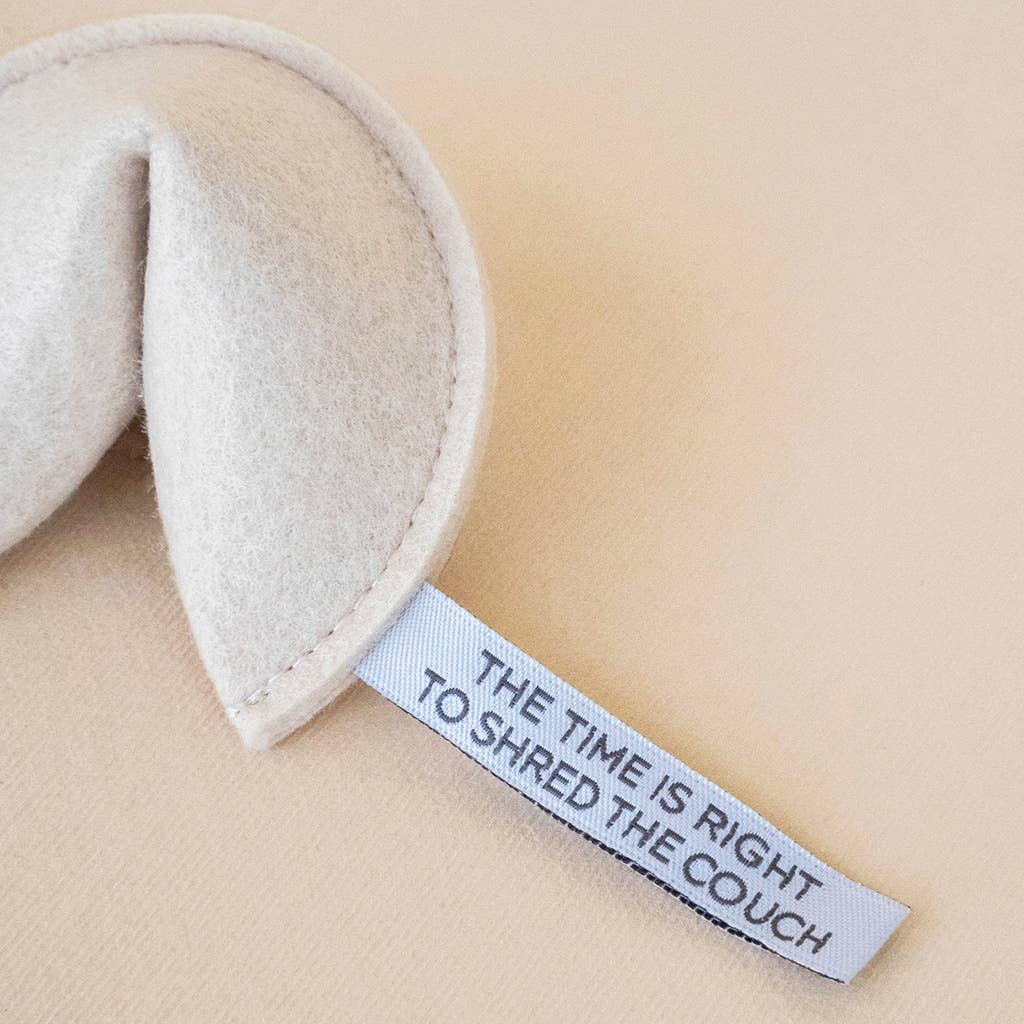 The MODERNBEAST - Kitty Fortune Cookie Organic Catnip Cat Toy sits on a light surface, its beige eco felt shaped like a fortune cookie with a paper fortune reading, "THE TIME IS RIGHT TO SHRED THE COUCH.