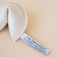 The MODERNBEAST - Kitty Fortune Cookie Organic Catnip Cat Toy sits on a light surface, its beige eco felt shaped like a fortune cookie with a paper fortune reading, "THE TIME IS RIGHT TO SHRED THE COUCH.