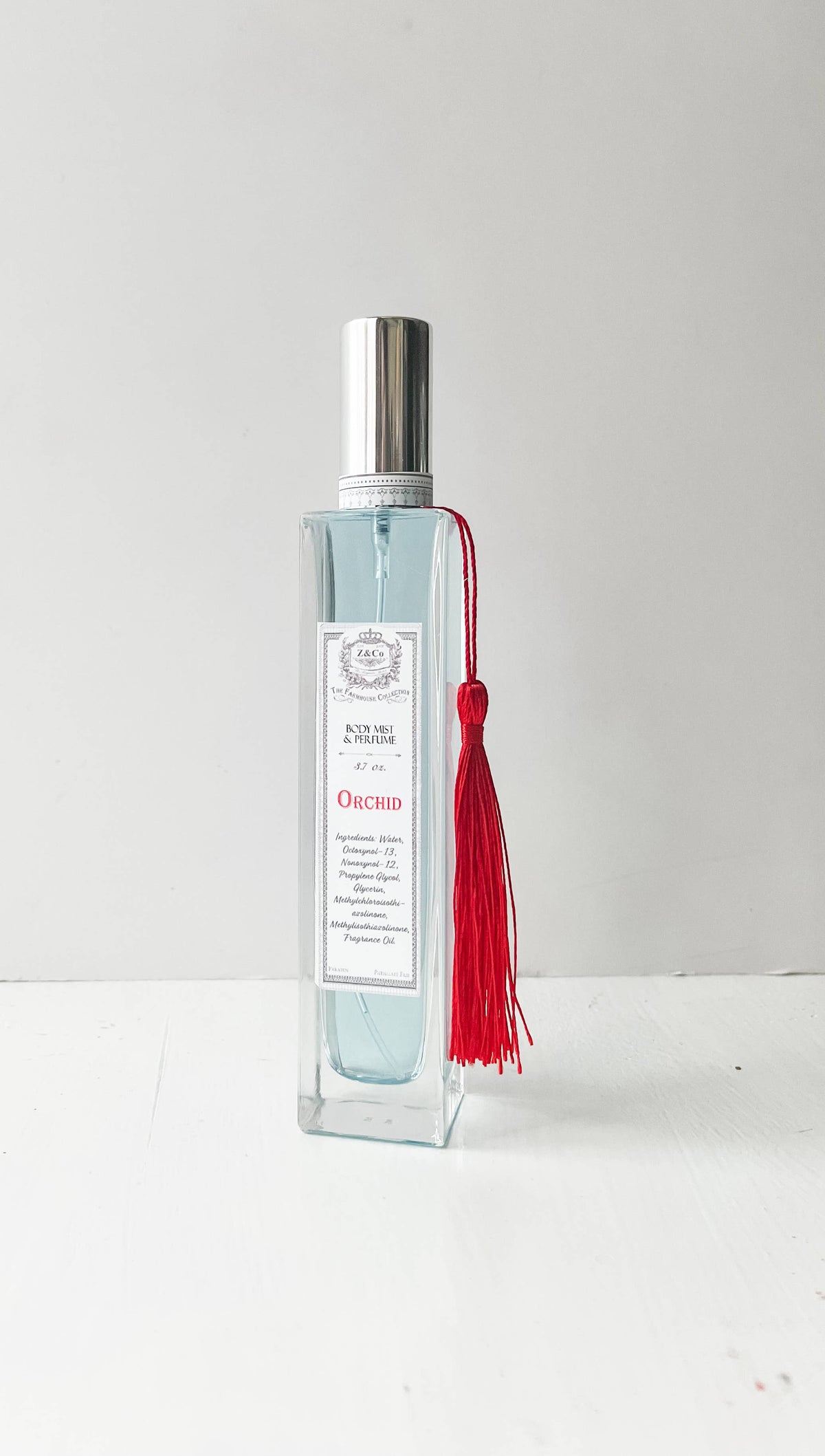A clear rectangular bottle of Z&Co. Farmhouse Body Mist - Orchid, featuring a silver cap and a decorative red tassel, stands on a white surface, highlighting the brand's exotic fragrance against a plain light background.