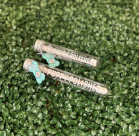 Two clear test tubes labeled "Wag & Bright Supply Co. - Puppy Polisher Eco Toothbrush - (Medium)" are placed on a lush green leafy surface. Each tube, crafted from eco-friendly wheat straw bioplastic, has a decorative blue tag attached to it.