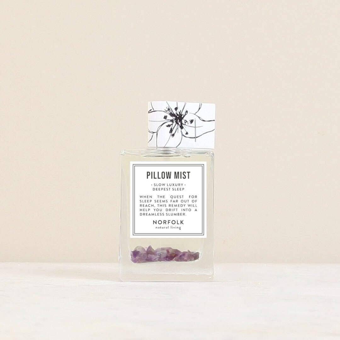 A clear glass bottle labeled "Norfolk Natural Living Deepest Sleep Pillow Mist" with small purple crystals sits on a light surface against a beige background. This lavender mist by Norfolk Natural Living promotes deep, restful sleep.