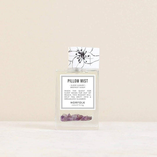 A clear glass bottle labeled "Norfolk Natural Living Deepest Sleep Pillow Mist" with small purple crystals sits on a light surface against a beige background. This lavender mist by Norfolk Natural Living promotes deep, restful sleep.
