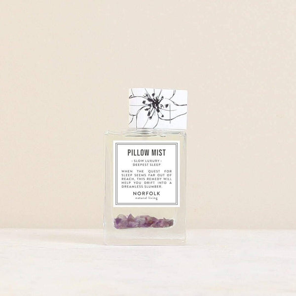 A clear glass bottle labeled "Norfolk Natural Living Deepest Sleep Pillow Mist" with small purple crystals sits on a light surface against a beige background. This lavender mist by Norfolk Natural Living promotes deep, restful sleep.