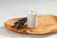The Sonoma Lavender Massage Candle by Sonoma Lavender rests in a simple holder on a wooden tray beside dried lavender, creating spa-like relaxation. The setup sits on a light, textured surface for a serene ambiance.