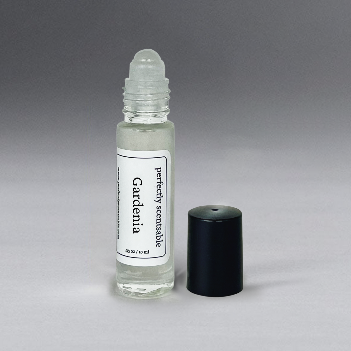 A small glass roller bottle containing a clear liquid labeled "Perfectly Scentsable Gardenia Perfume Oil - Rollerball." The bottle holds a long-lasting aroma of gardenia perfume oil and features a rollerball applicator at the top. A black cap rests next to it on a light grey surface, set against a gradient background that transitions from light to dark grey.