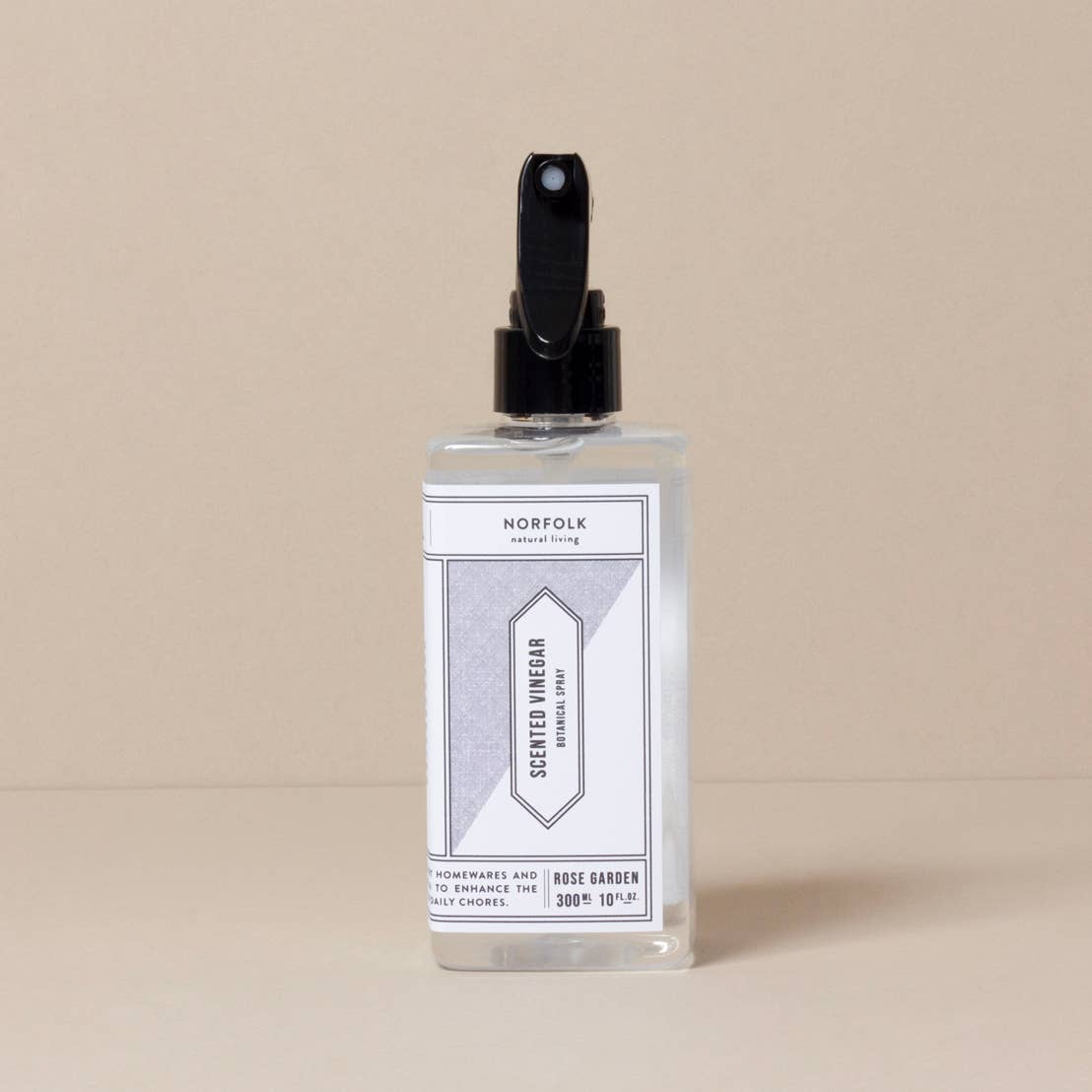 A clear spray bottle of Norfolk Natural Living Rose Garden Scented Vinegar, with a minimalist design and black nozzle, offers a natural cleaner with a rose garden fragrance. The product is set against a plain beige background.