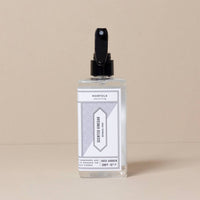 A clear spray bottle of Norfolk Natural Living Rose Garden Scented Vinegar, with a minimalist design and black nozzle, offers a natural cleaner with a rose garden fragrance. The product is set against a plain beige background.