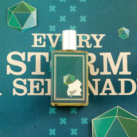 A bottle of Imaginary Authors Every Storm a Serenade - Large Size perfume, featuring a green and white geometric design on its label, sits on a blue-green surface. The background reveals partial text that reads "EVERY STORM" alongside abstract polygon shapes in varying shades of green, evoking the scents of eucalyptus and Danish spruce.