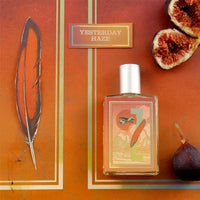 A large-sized bottle of "Imaginary Authors Yesterday Haze" perfume is displayed on a colorful background featuring a feather illustration, sliced figs, and an additional whole fig. The 50ml bottle showcases earthy tones, complemented by a silver cap, exuding a warm and rustic aesthetic. This seductive fragrance offers an inviting allure.