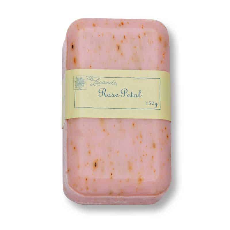 A rectangular bar of pink soap with small brown flecks, labeled "La Lavande Joie de Vivre Rose Petal Hand, Face and Body Soap 150g," wrapped in a cream-colored paper band. The soap, featuring a smooth surface and appearing to be handmade or artisanal, is crafted using the triple milling process for enhanced quality.