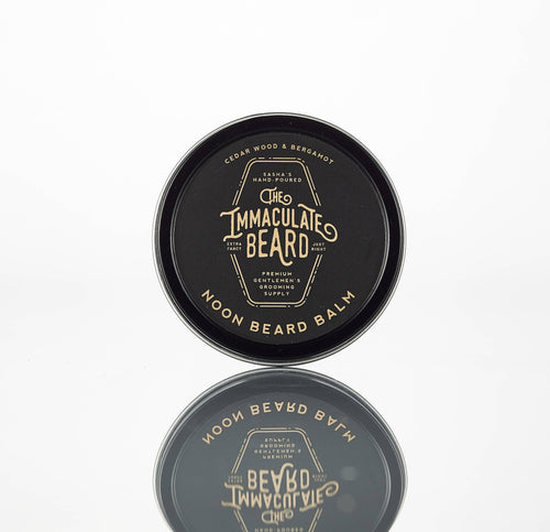 A round black tin of The Immaculate Beard NOON Beard Balm with cedarwood bergamot scent and natural ingredients stands upright on a white background, its reflection visible below.