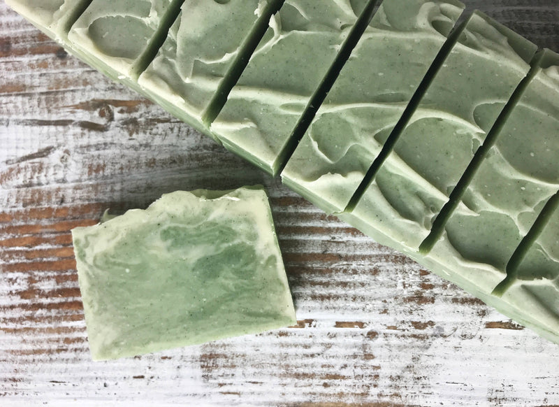 A rustic wooden surface is covered with pale green Wild Botanicals Eucalyptus Cajeput Spirulina Soap bars. One soap bar is placed separately below the orderly stack of six bars. Made with organic ingredients, the soap has a textured, wavy pattern on top, displaying a handmade appearance and eco-friendly packaging from Wild Botanicals.