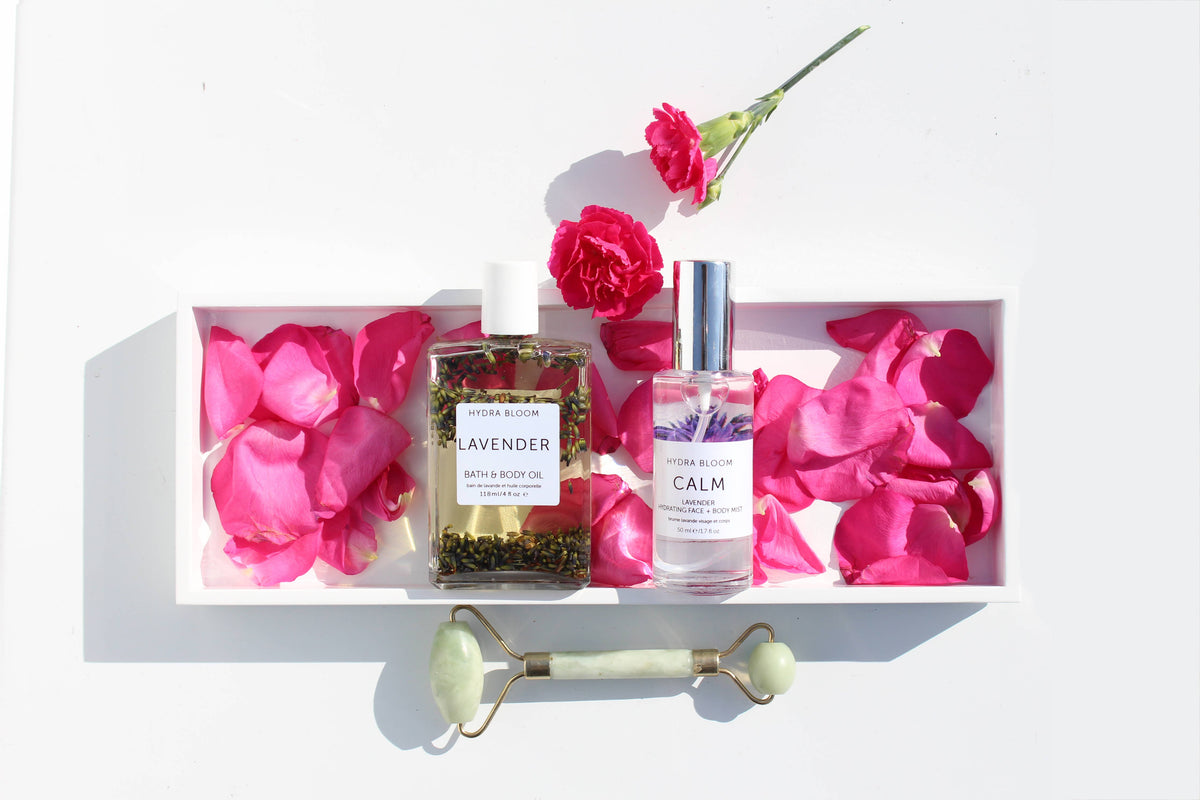 A collection of spa essentials including a box of Hydra Bloom Beauty Luxe Lavender Bath and Body Oil, a calm aromatherapy spray, and a jade roller surrounded by pink rose petals and an organic bath oil on a white background.