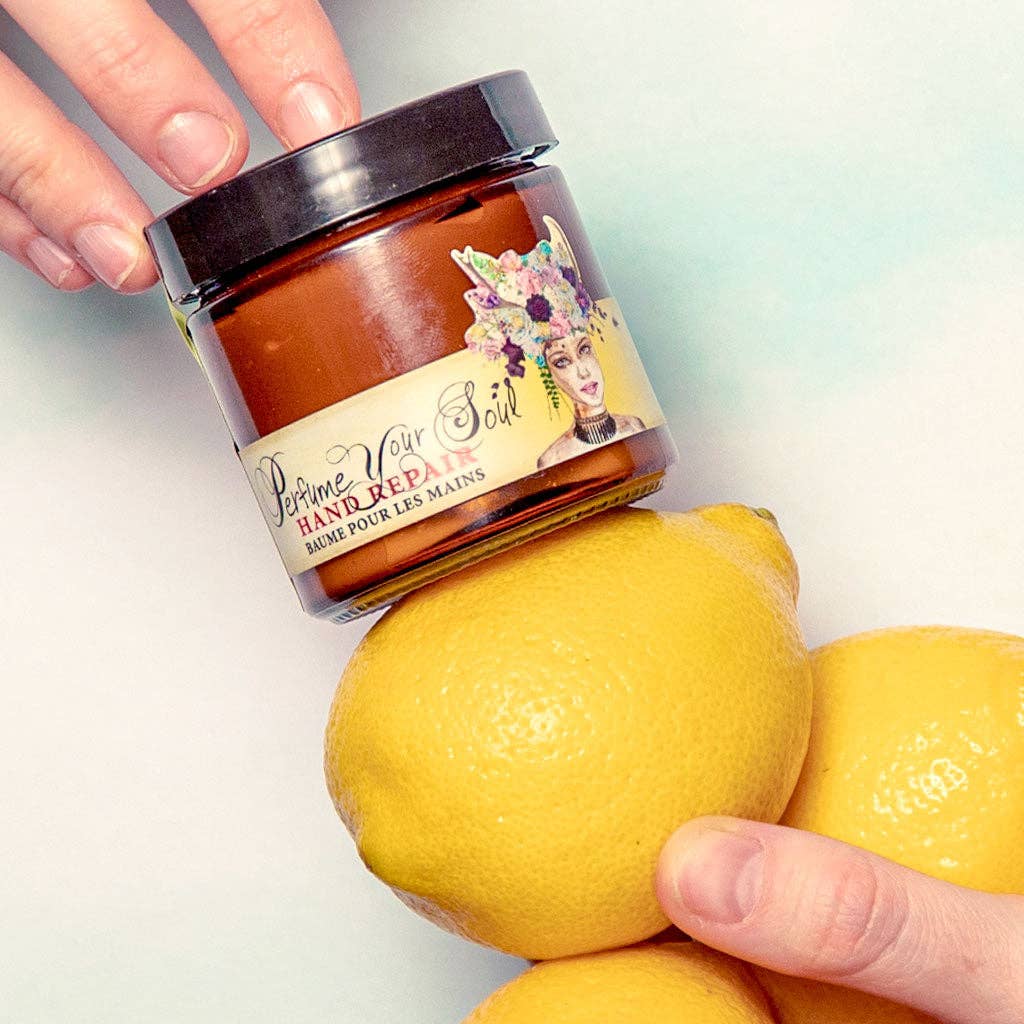 A jar of Barefoot Venus Lemon Freckle Hand Repair Balm is held above two yellow lemons, with one hand holding the jar and another holding a lemon against a light background.