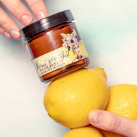 A jar of Barefoot Venus Lemon Freckle Hand Repair Balm is held above two yellow lemons, with one hand holding the jar and another holding a lemon against a light background.