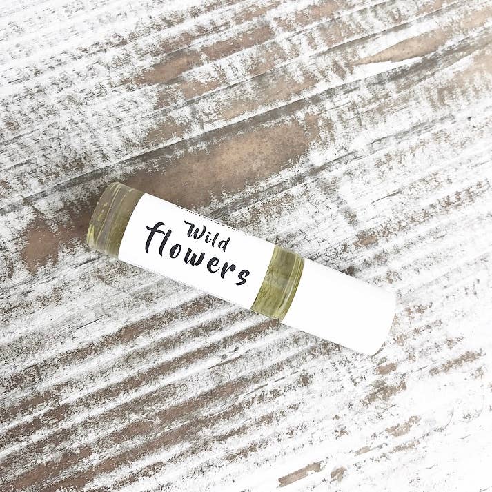 A small bottle labeled "Wild Botanicals Wildflowers Aromatherapy Roll On" with a white cap lies on a rustic, weathered wooden surface. The bottle appears to contain a clear or light-colored essential oil.