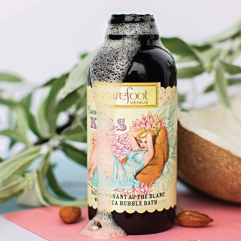 A bottle labeled "Barefoot Venus" featuring a vintage-style illustration of a relaxed woman surrounded by flowers, with foam overflowing from the top. Highlighting the Barefoot Venus Coconut Kiss Bubble Bath, the bottle is set against a backdrop of green leaves, a pink surface, and almonds.