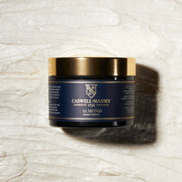 A black and gold jar of Caswell Massey Caswell~Massey Almond Shave Cream rests on a textured, creamy white surface with a rightward shadow; the product label is clearly visible.