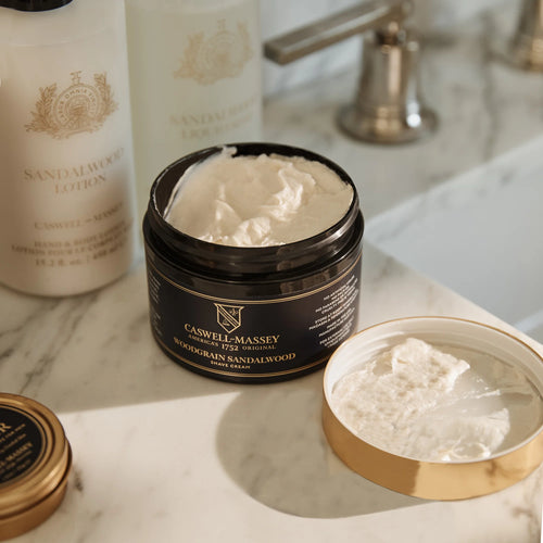 An open jar of Caswell-Massey Woodgrain Sandalwood Shave Cream sits on a marble counter with its lid, matching Sandalwood lotion and soap, and a metal faucet in the background. This cream by Caswell Massey provides a smooth, hydrating shave.
