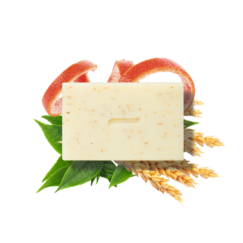 The Caswell-Massey Body Scrub Bar by Caswell Massey, featuring a minimal groove design, rests on green leaves, wheat stalks, and grapefruit peels. Made with natural ingredients, this beige speckled exfoliating soap is shown on a white background.