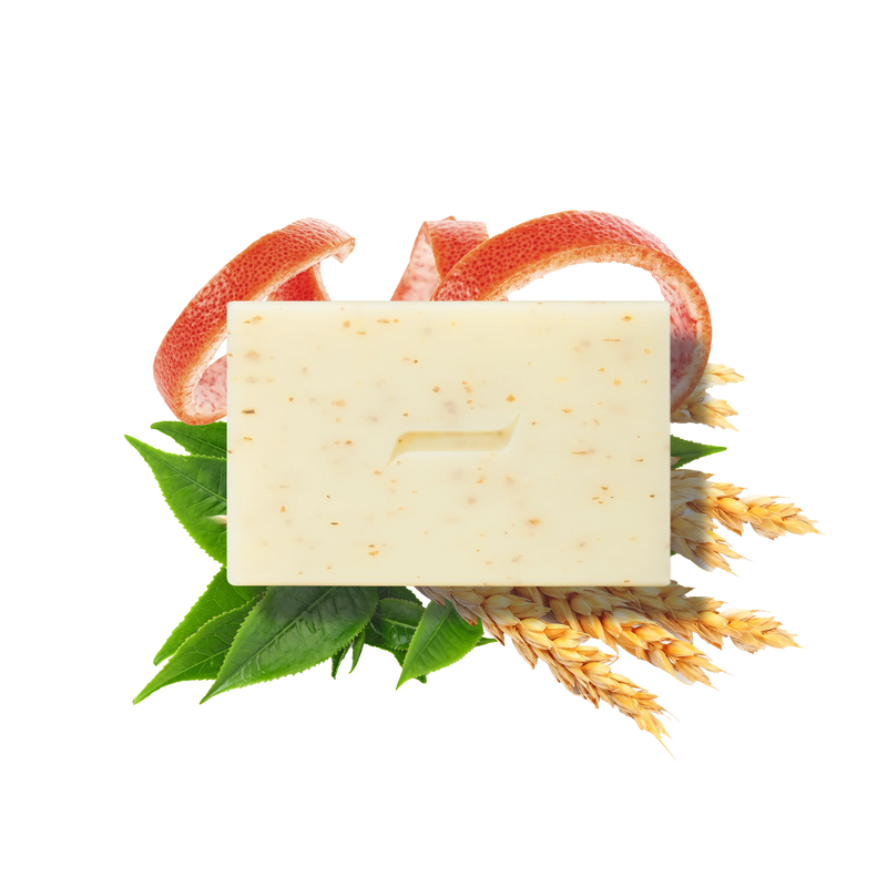 The Caswell-Massey Body Scrub Bar by Caswell Massey, featuring a minimal groove design, rests on green leaves, wheat stalks, and grapefruit peels. Made with natural ingredients, this beige speckled exfoliating soap is shown on a white background.