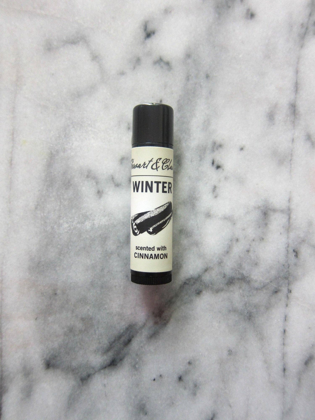 A tube of Stewart & Claire Winter Lip Balm with Cinnamon rests on a gray and white marbled surface, its label adorned with an illustration of a cinnamon stick.