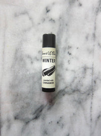 A tube of Stewart & Claire Winter Lip Balm with Cinnamon rests on a gray and white marbled surface, its label adorned with an illustration of a cinnamon stick.